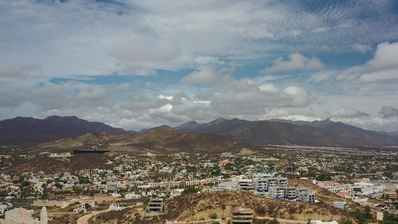 Aerial view one second into the Los Cabos clip, with white buildings in the lower frame, brown hills in the middle distance, mountains beyond, and no visible people.