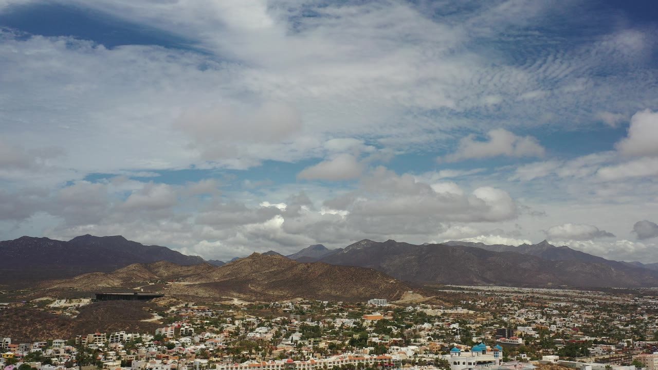 Opening frame of Los Cabos, Mexico from above, with white buildings at the bottom edge, brown hills and dark mountains on the horizon, a large cloud-filled sky, and no visible people.
