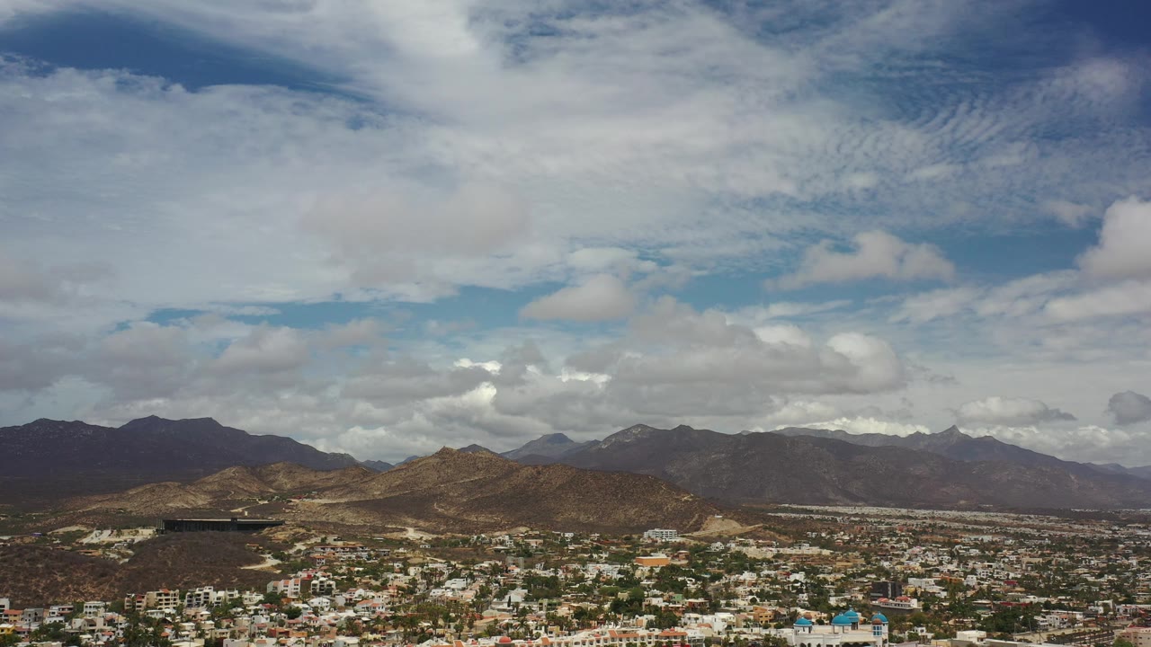 Wide view over Los Cabos buildings toward hills and a bright cloudy sky.