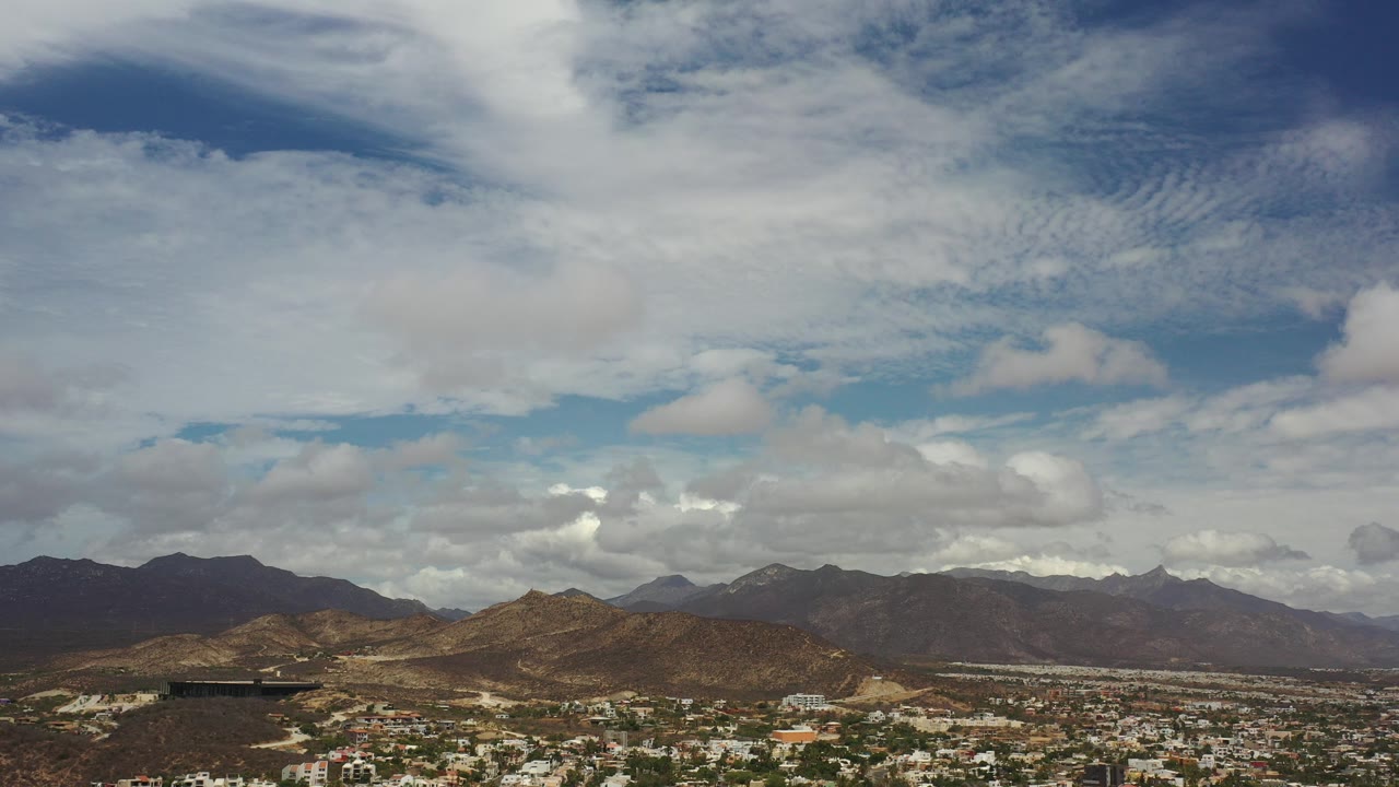 Wide aerial view in Los Cabos, Mexico, with blue sky, thick white clouds, brown hills, distant mountains, and a low city strip along the bottom, no visible people.