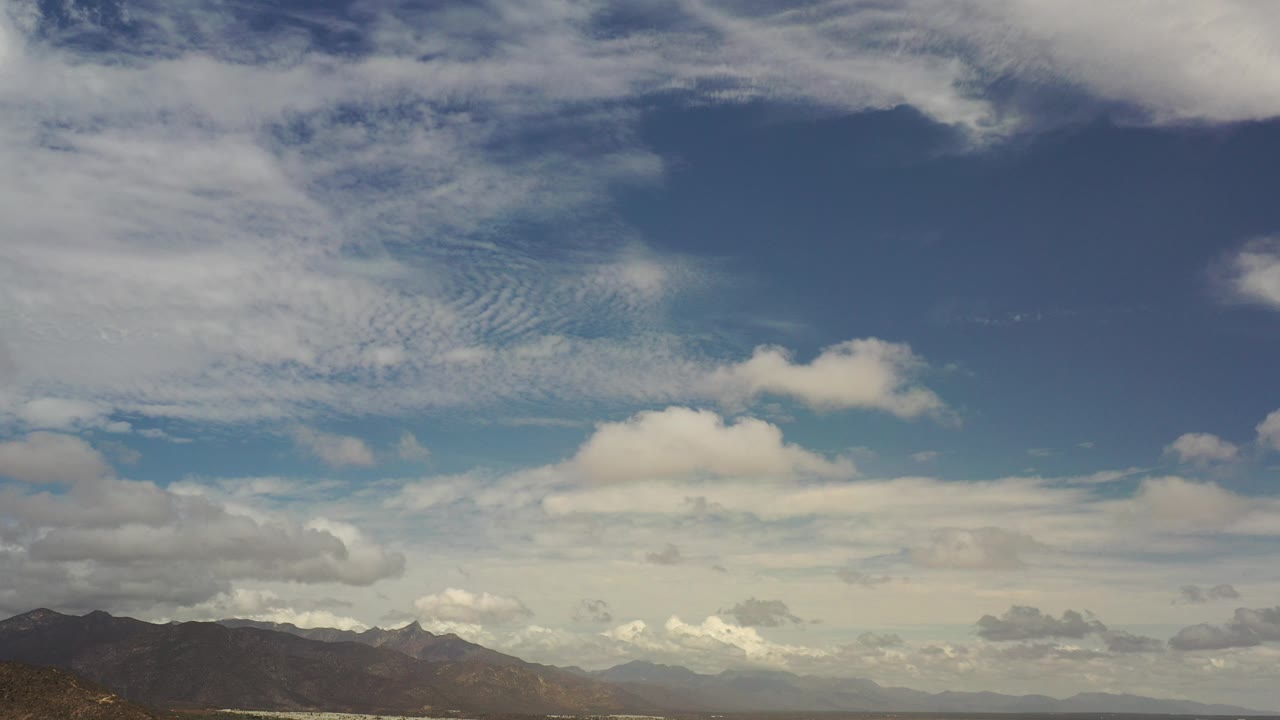 Wide Los Cabos, Mexico view with layered clouds dominating the blue sky, dark mountain ridges along the lower edge, and no visible people.