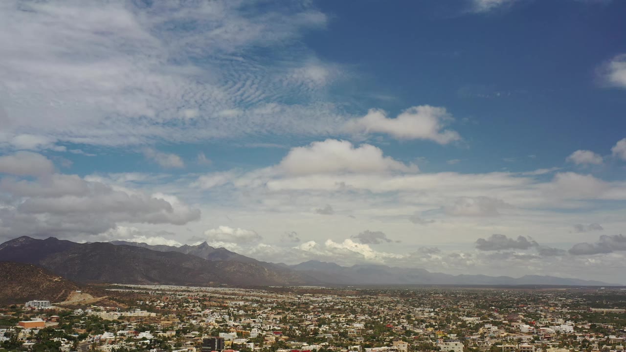 Aerial frame over Los Cabos, Mexico, with rooftops and streets filling the lower edge, a mountain ridge across the horizon, and white clouds spread through the sky, with no visible people.