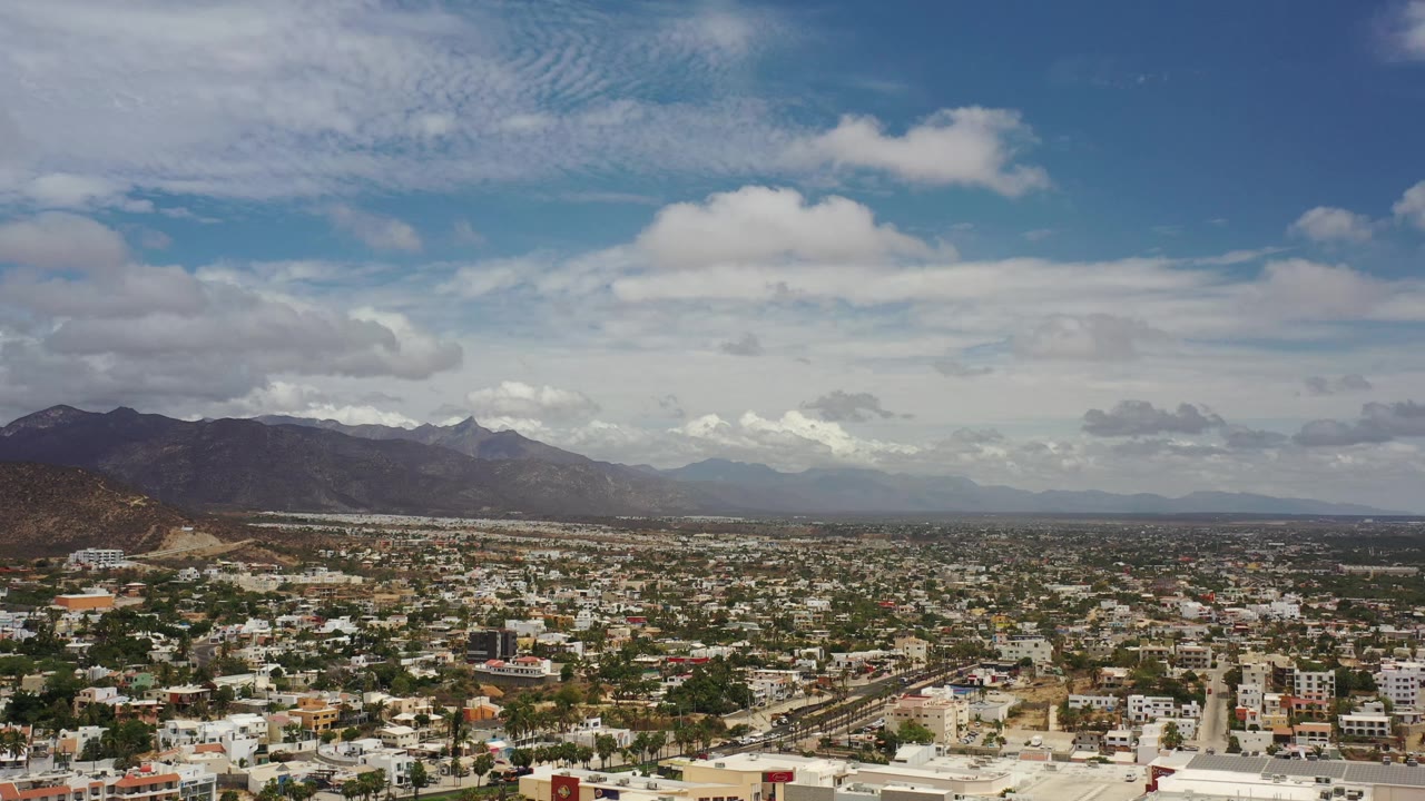 Los Cabos appears as a dense low-rise townscape beneath a broad cloud layer and distant mountain horizon, with no visible people.