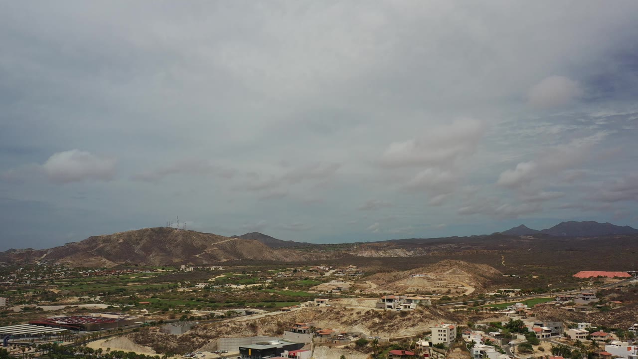 Wide aerial view of hilly plain, scattered buildings, and cloudy sky.