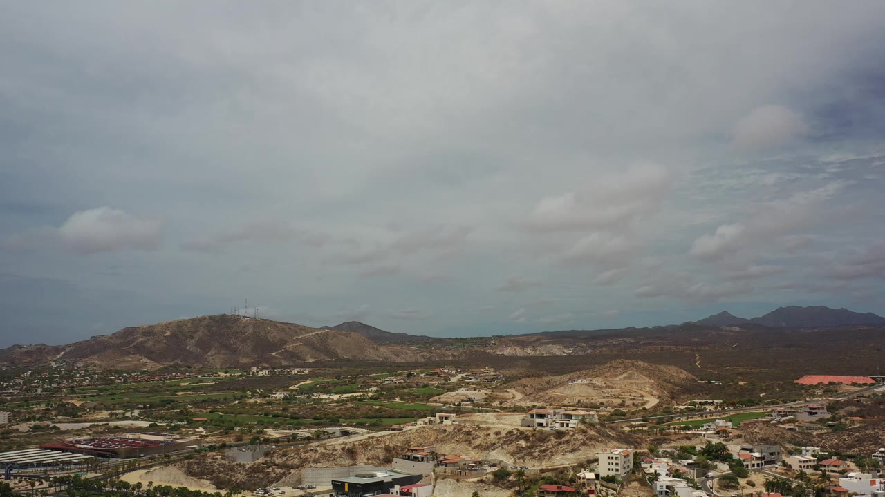 Los Cabos aerial frame with low brown hills in the foreground, a developed plain in the midground, dark distant ridges, and no visible people.