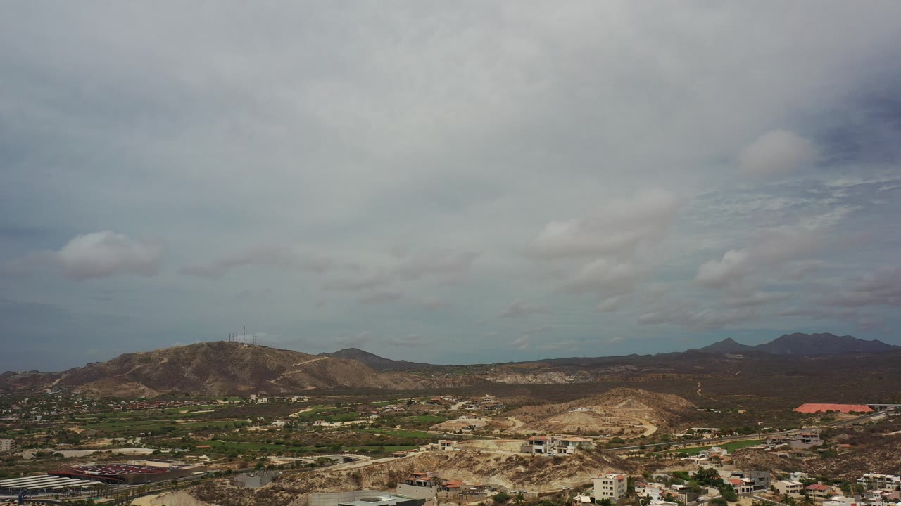 Wide aerial frame over Los Cabos, Mexico with brown hills, green scrub patches, scattered buildings, pale roads, and no visible people.