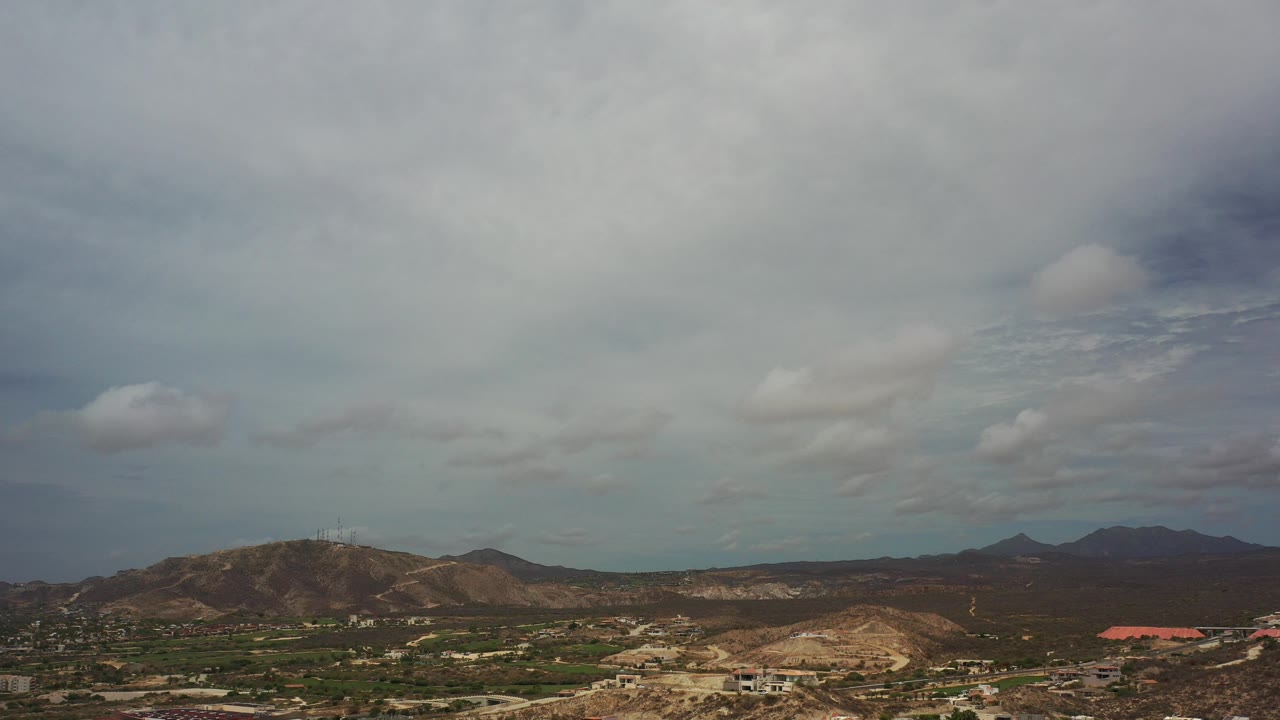 Wide aerial view over Los Cabos, Mexico, with low hills, green patches, distant dark mountains, and a cloud-filled sky, with no visible people.