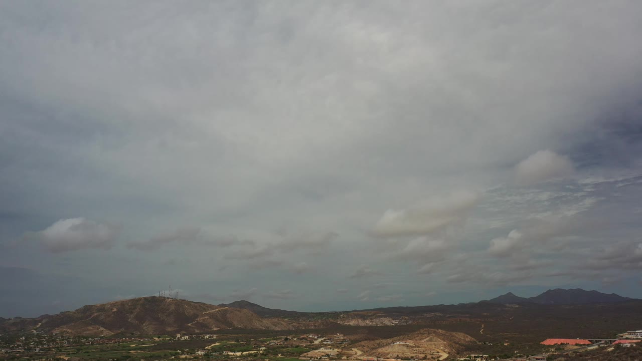 Los Cabos hills and distant dark peaks sit below a broad ceiling of gray clouds, with no visible people in the landscape.