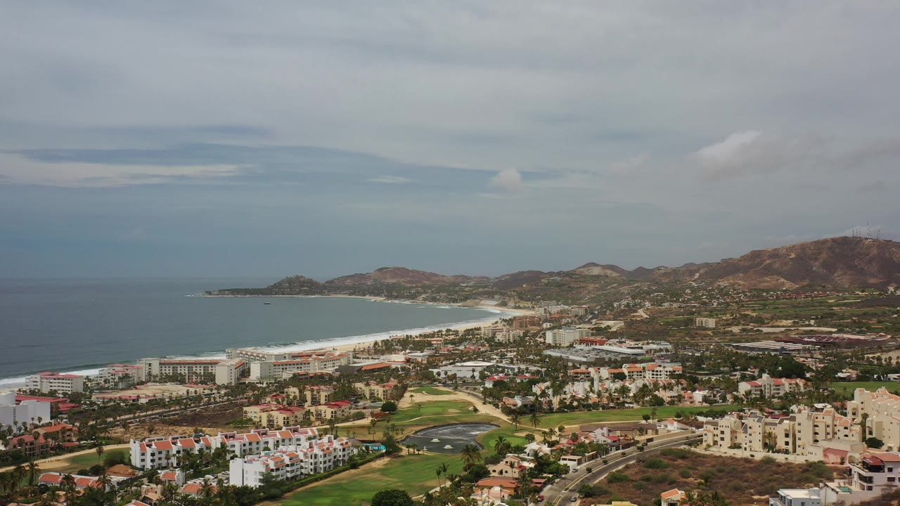 Aerial view of coastal town, ocean, beach, buildings, green grass, and mountains.