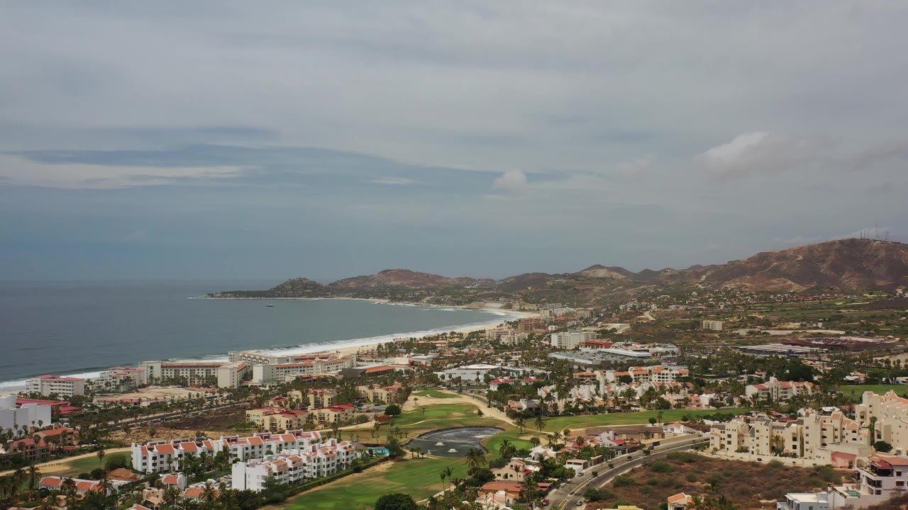 Aerial view of Los Cabos, Mexico with the shoreline running across the frame, white waves along the beach, clustered buildings, roads, green grounds, and no visible people.
