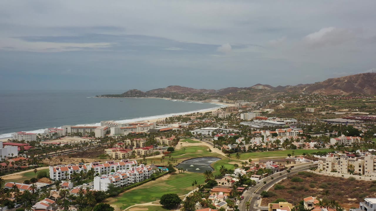 Wide aerial frame in Los Cabos, Mexico, showing the coastline slightly farther across the frame, green fairways and water hazards below, clustered buildings, hills, and no discernible people.