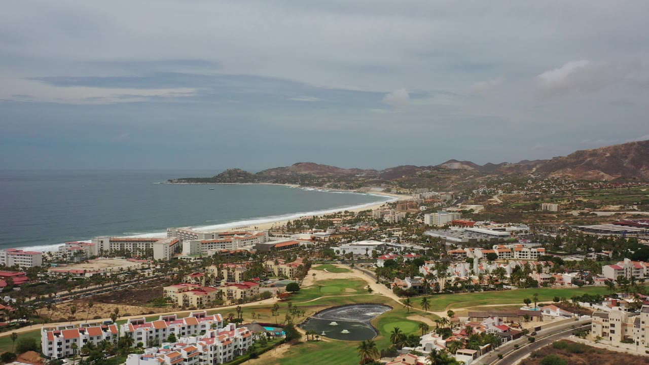 Opening wide aerial frame over Los Cabos, Mexico with ocean on the left, a pale beach curving through the midground, city buildings and golf greens below, mountains behind, and no visible people.