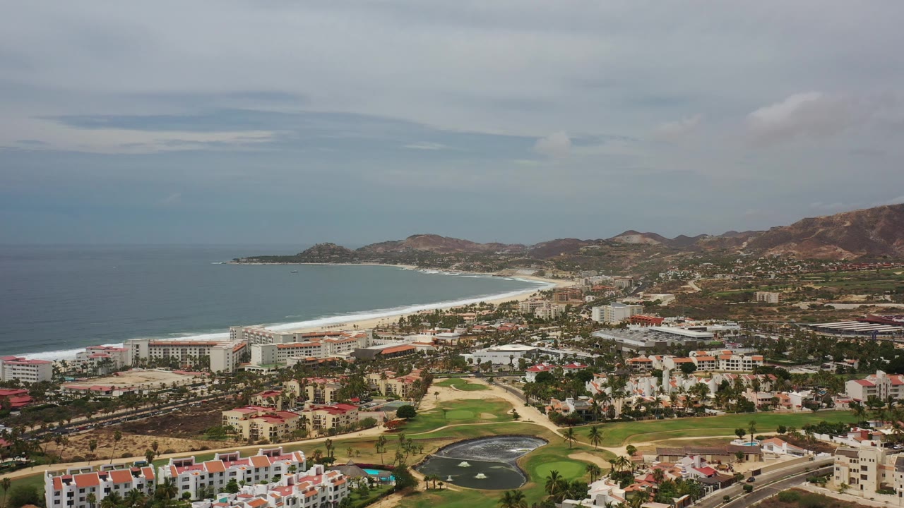High view of Los Cabos beach district, ocean bay, golf landscaping, buildings, and hills.
