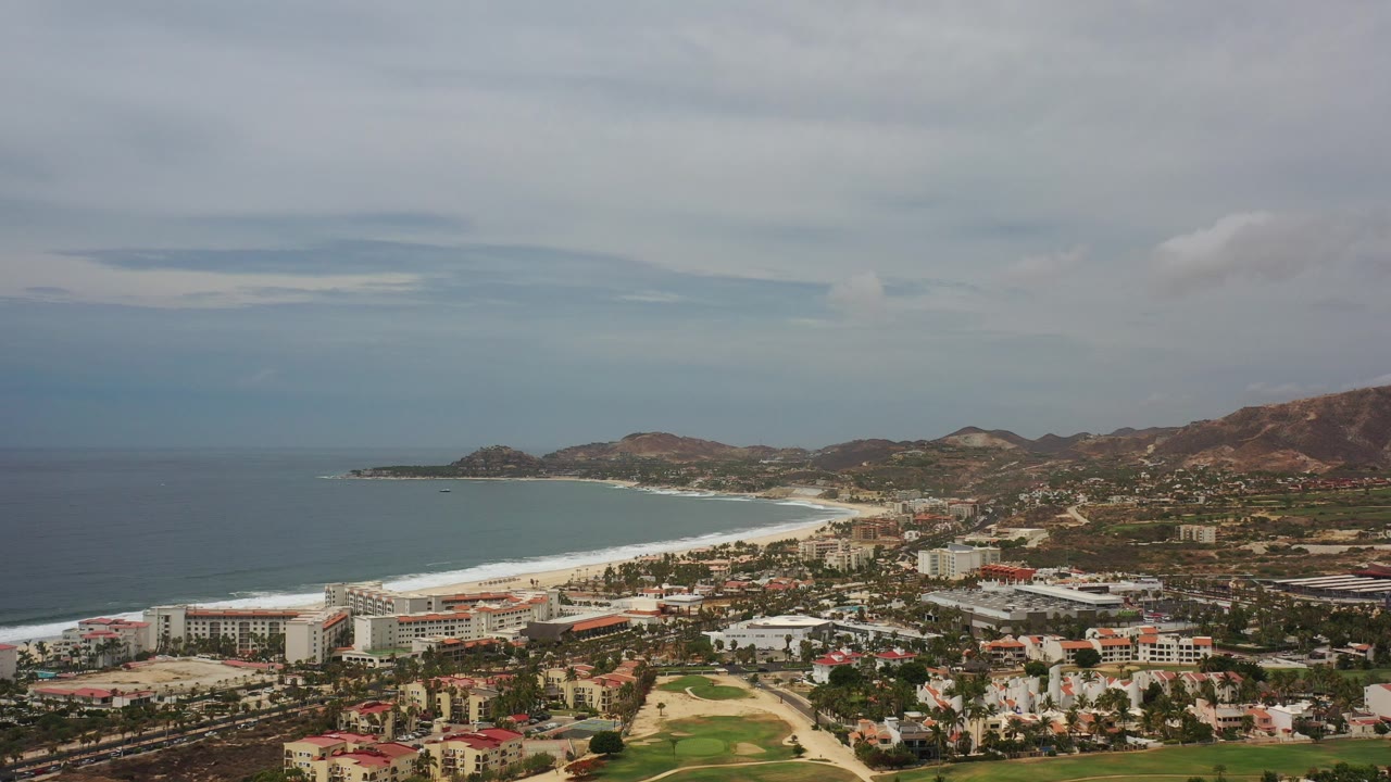 Wide aerial frame over Los Cabos, Mexico, showing blue ocean, a pale sandy beach, low coastal buildings, hills in the distance, and no visible people.