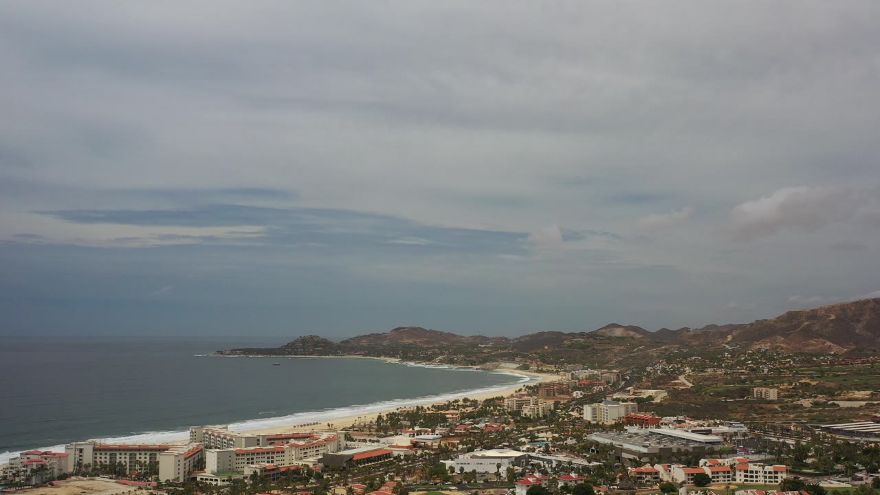 Opening aerial view over Los Cabos, Mexico, with dark ocean at left, a pale surf line along the beach, town buildings below, brown hills beyond, and no visible people.