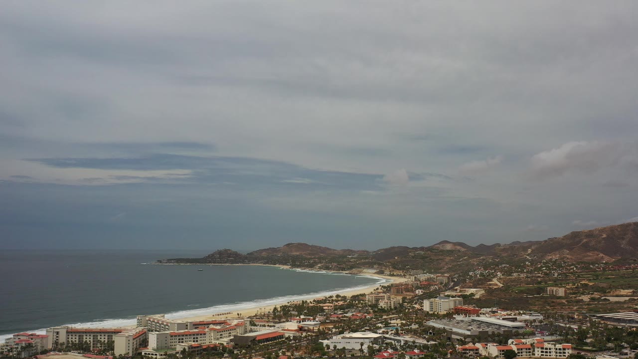 Cloudy aerial coastline with ocean, beach, town, and hills.