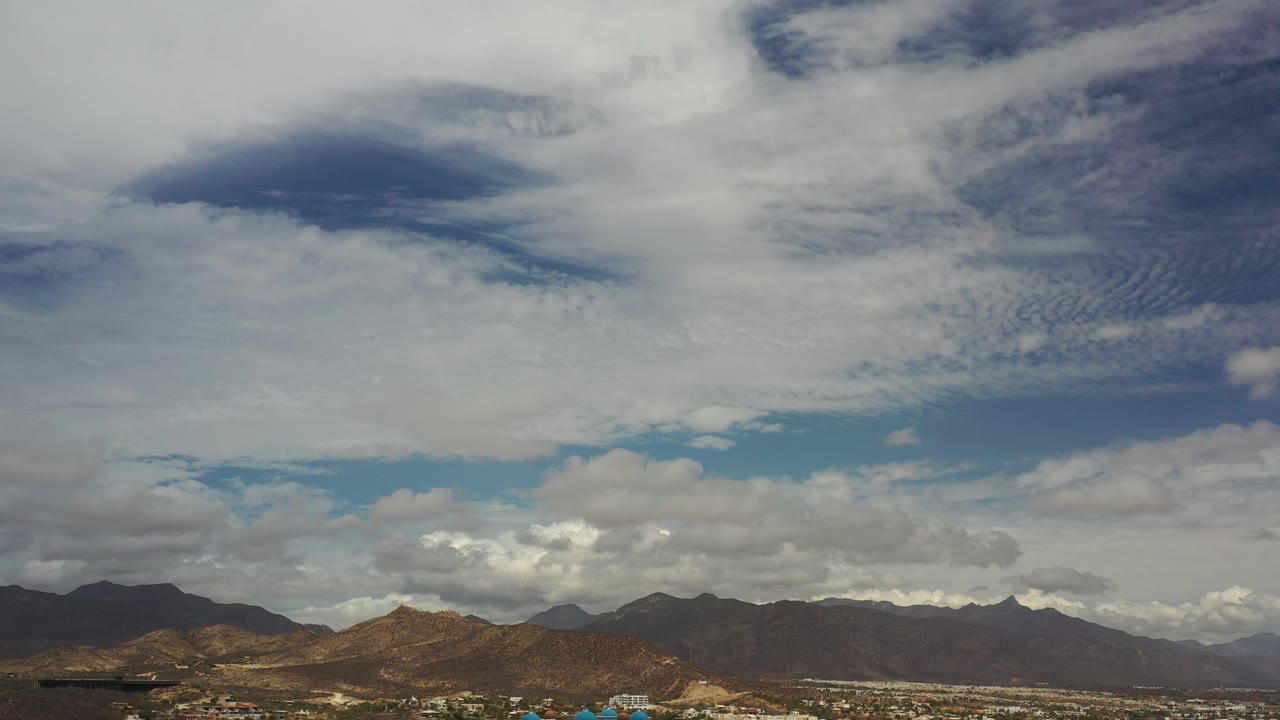 Opening frame of Los Cabos, Mexico with broad white and gray clouds filling the sky above dark mountain ridges and no visible people.