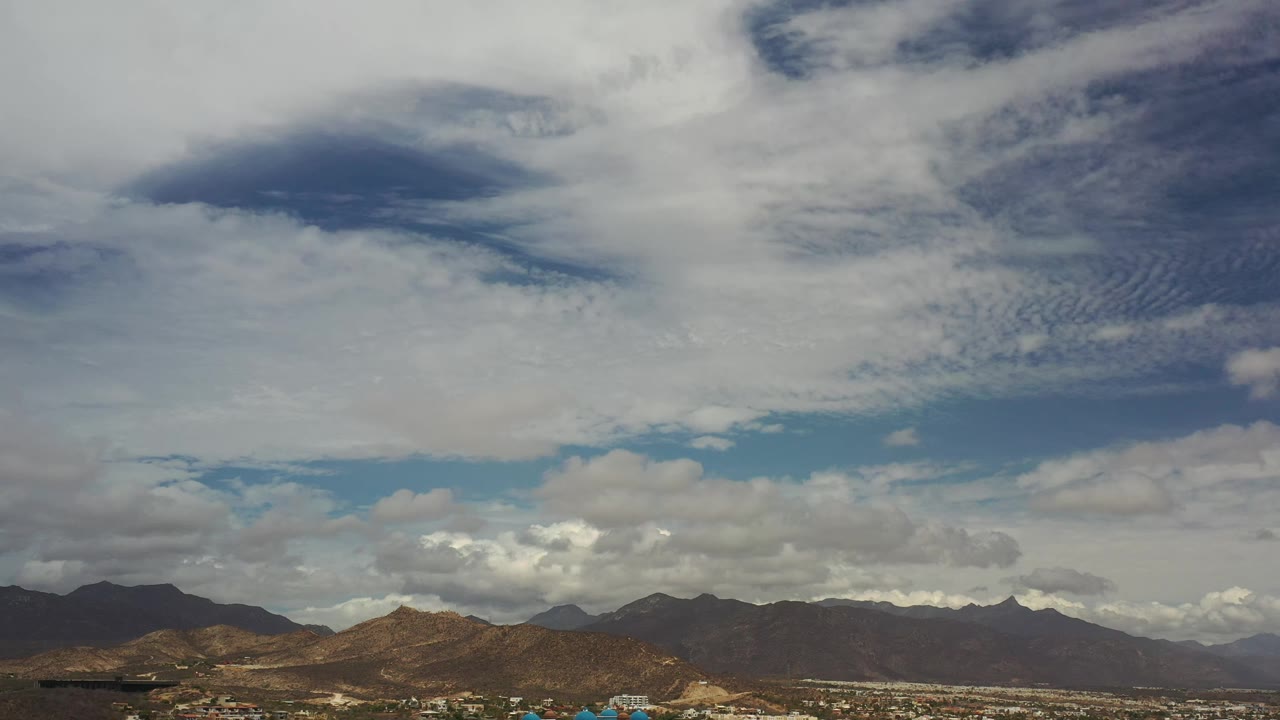 A similar Los Cabos mountain view one second in, with a darker cloud patch near the upper left and a low line of ridges beneath the sky, no visible people.