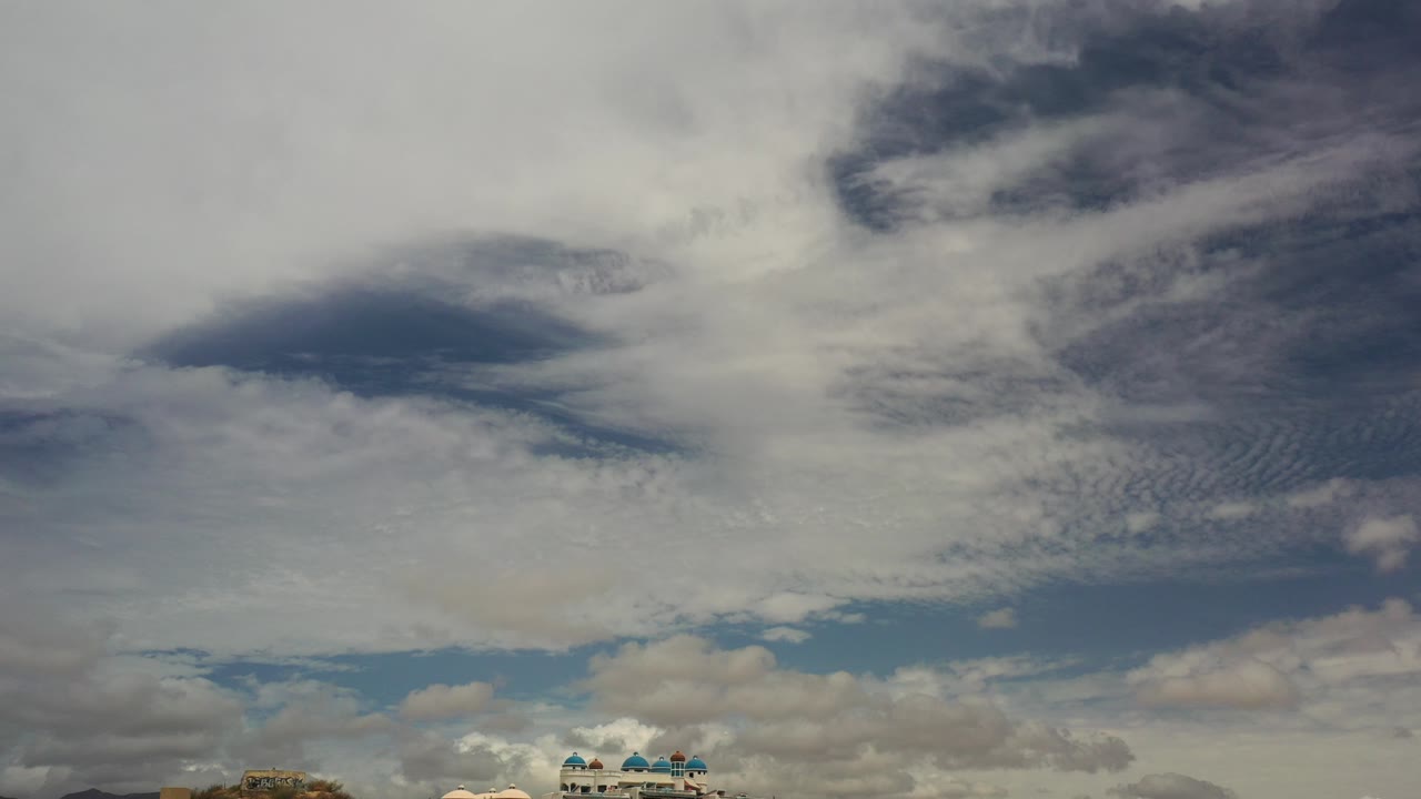 Opening frame of the Los Cabos clip showing layered white and gray clouds across a blue sky, a thin strip of rooftops and hillside near the bottom, and no visible people.