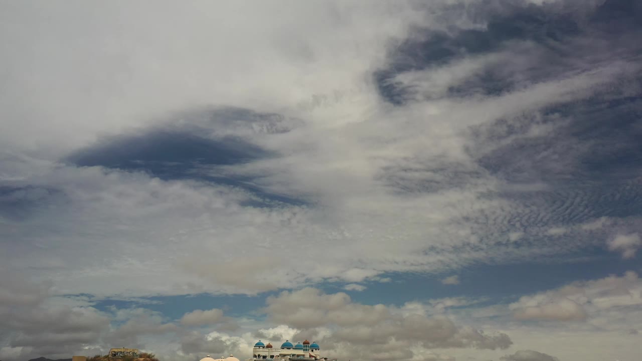 Los Cabos sky view with broad white cloud cover, darker blue-gray patches, small distant buildings along the bottom edge, and no visible people.