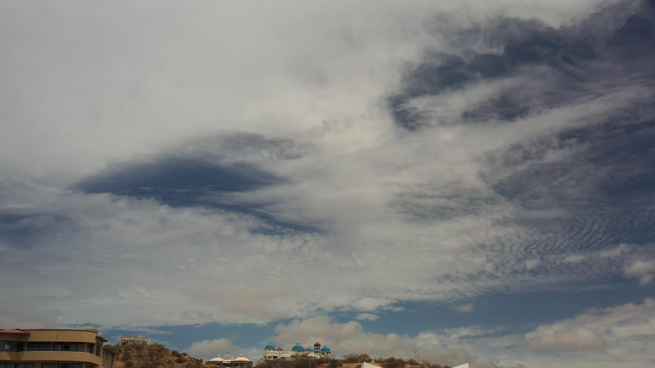 Wide Los Cabos frame with layered clouds and blue sky over low hillside buildings, with no visible people.