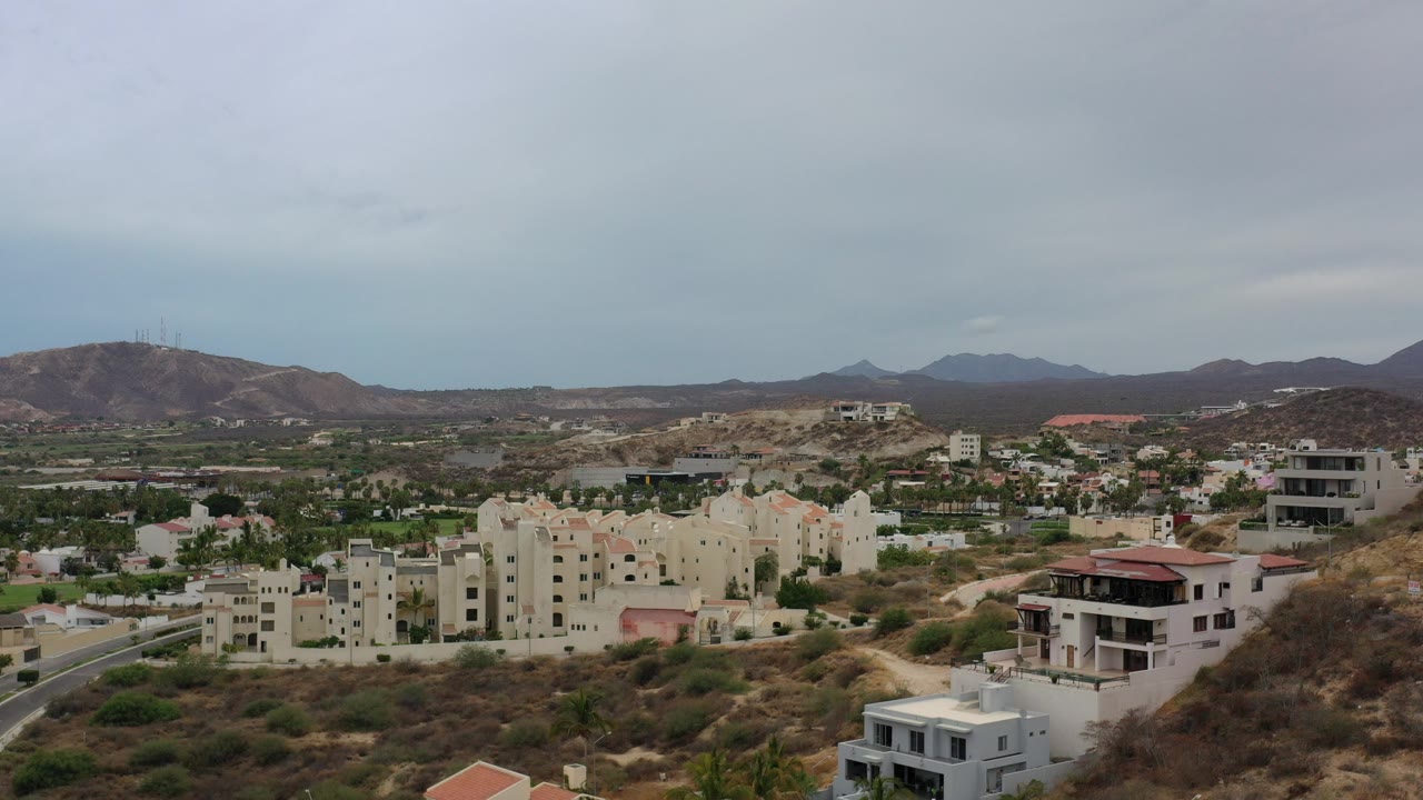 Opening aerial frame over Los Cabos, Mexico, showing cream residential buildings, scattered hillside homes, brown slopes, distant mountains, and no visible people.