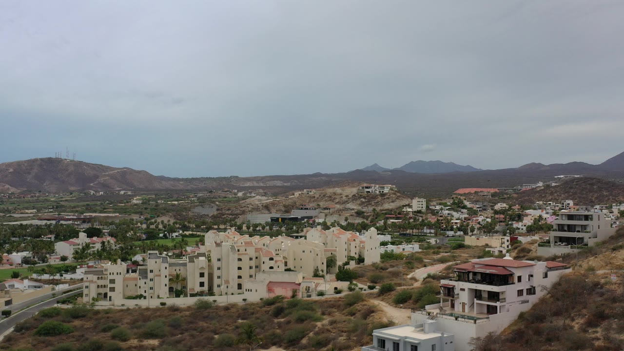 Drone view of cream buildings below brown hills and distant ridges.