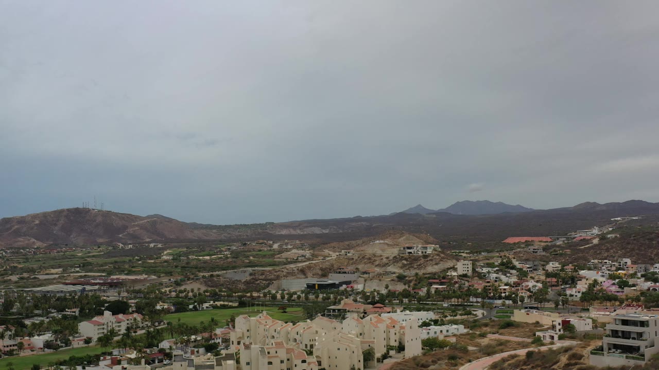 Los Cabos aerial frame showing more of the valley floor, roads, pale buildings, green lawns, brown hills, and no visible people.