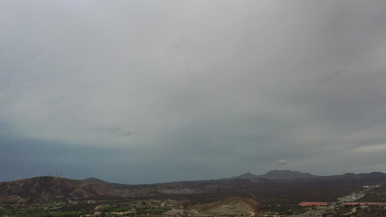 Wide Los Cabos landscape with gray cloud cover, low desert hills, distant mountain ridges, small buildings near the bottom edge, and no visible people.