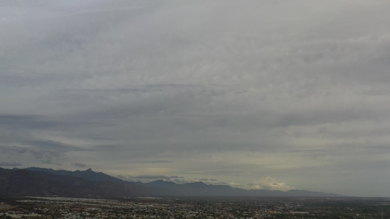 Opening aerial frame of Los Cabos with low city buildings across the bottom, distant dark mountains, an overcast sky, and no visible people.