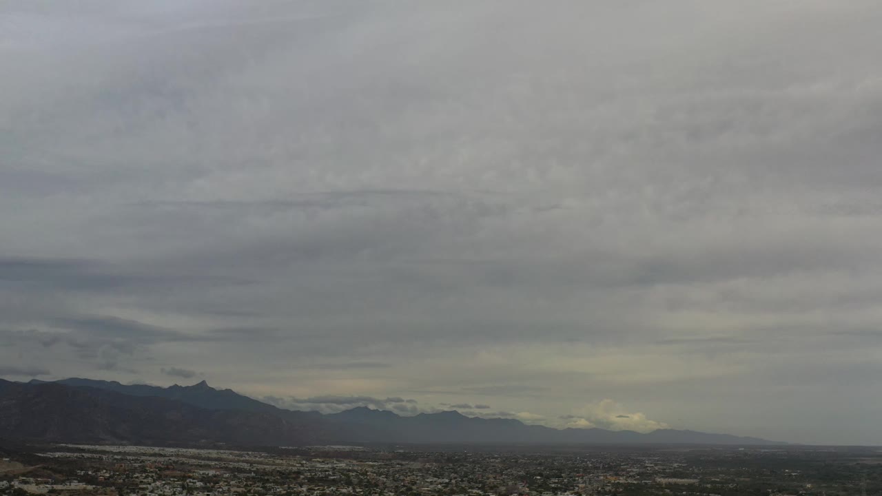 Los Cabos aerial view one second in, showing the urban area lower in frame, layered mountains on the horizon, thick gray clouds, and no visible people.