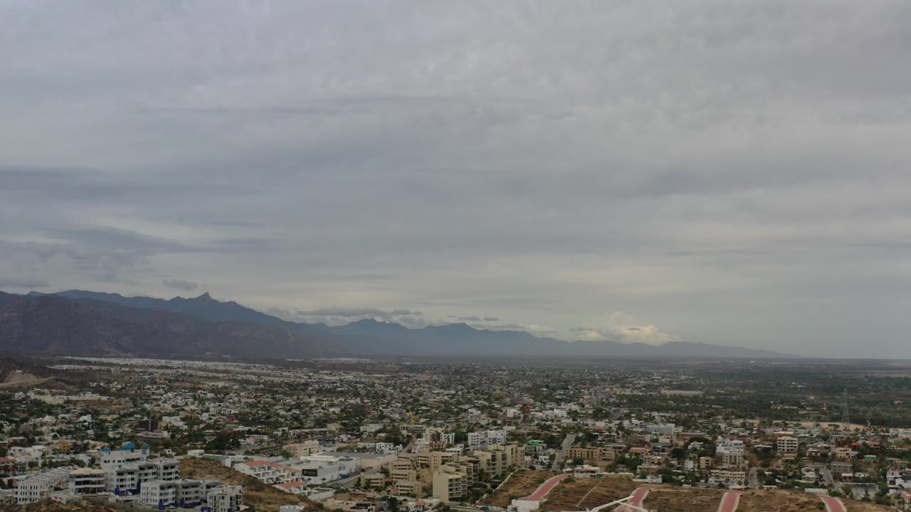 Wide aerial city skyline beneath gray clouds and distant mountains.