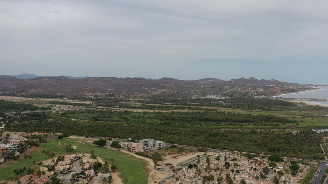 Opening aerial frame over Los Cabos, Mexico, showing green valley terrain, scattered buildings, distant brown hills, pale coastal water at right, and no visible people.