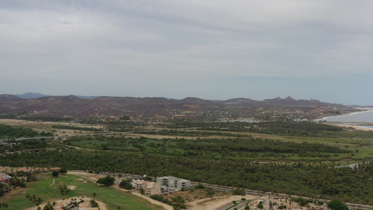 Opening frame of a wide Los Cabos, Mexico aerial view with green vegetation, distant brown hills, a pale coastline, overcast sky, and no visible people.