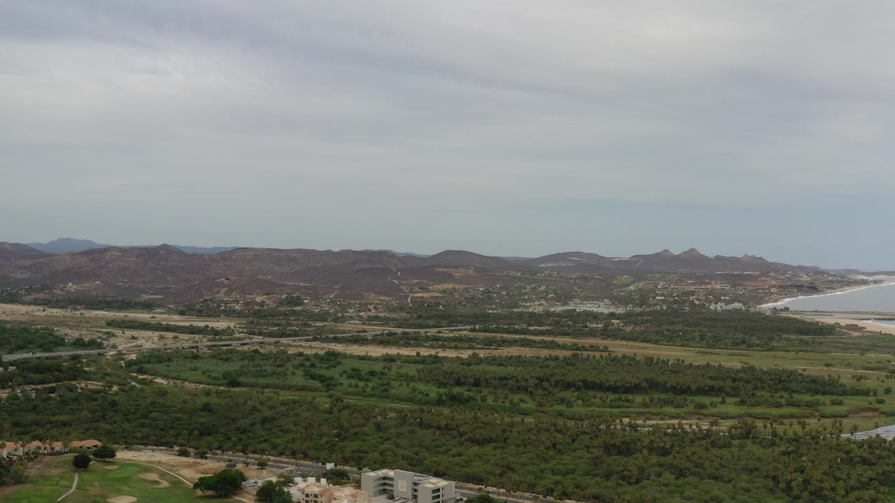 Opening frame of Los Cabos, Mexico with green valley land below brown hills, a pale coastline on the right, cloudy sky above, and no visible people.