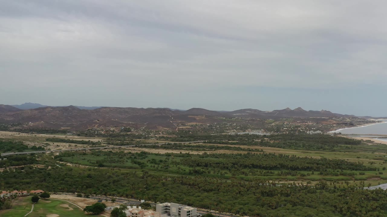 Early frame from Los Cabos, Mexico showing layered green fields, dry hills across the horizon, coastline at the right edge, gray cloud cover, and no visible people.
