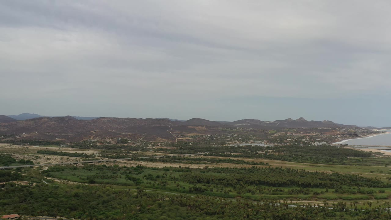 Second frame of Los Cabos, Mexico showing the same elevated landscape with the bay more centered at right, layered green fields, brown hills, cloudy sky, and no visible people.