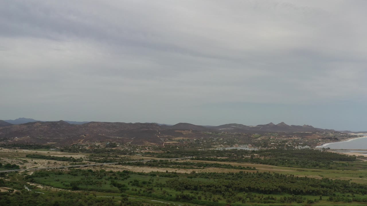 Opening frame of a wide Los Cabos aerial landscape with green foreground fields, low brown hills, a pale ocean shoreline at the right edge, gray clouds, and no visible people.
