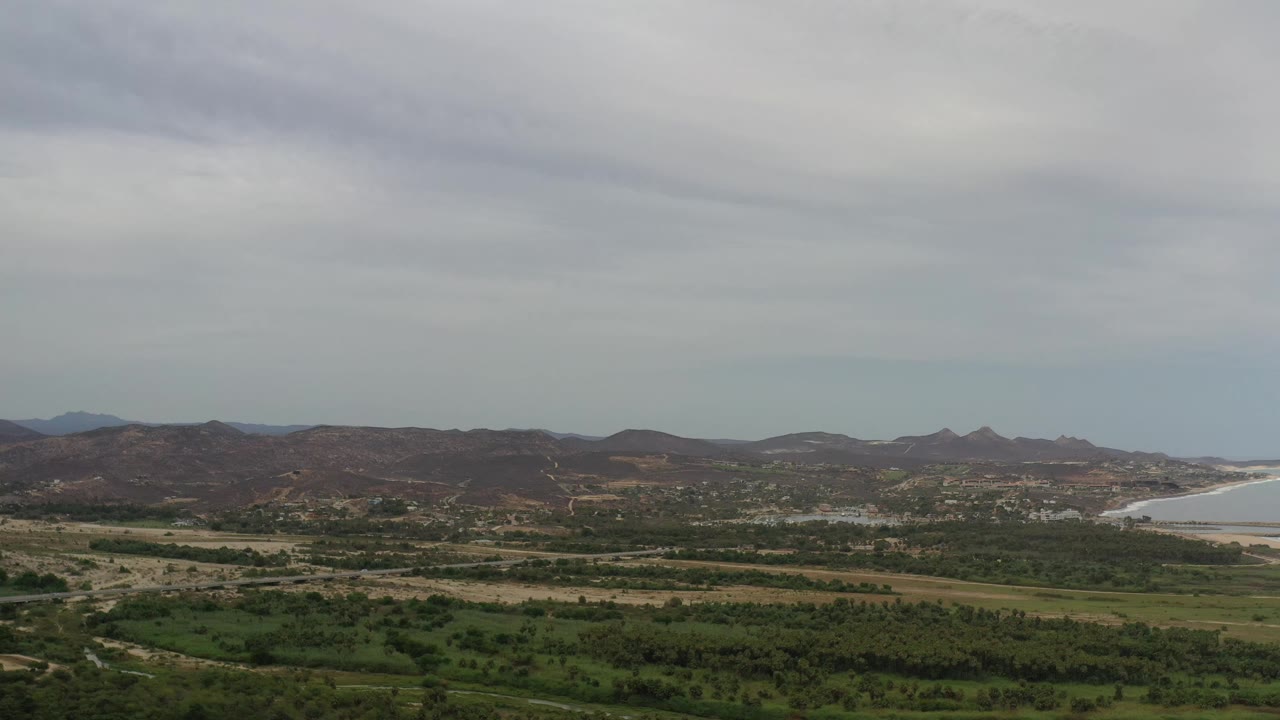 At 0:01, the Los Cabos view holds on layered green fields and scrub, brown ridges across the horizon, a light-toned ocean curve to the right, and no visible people.