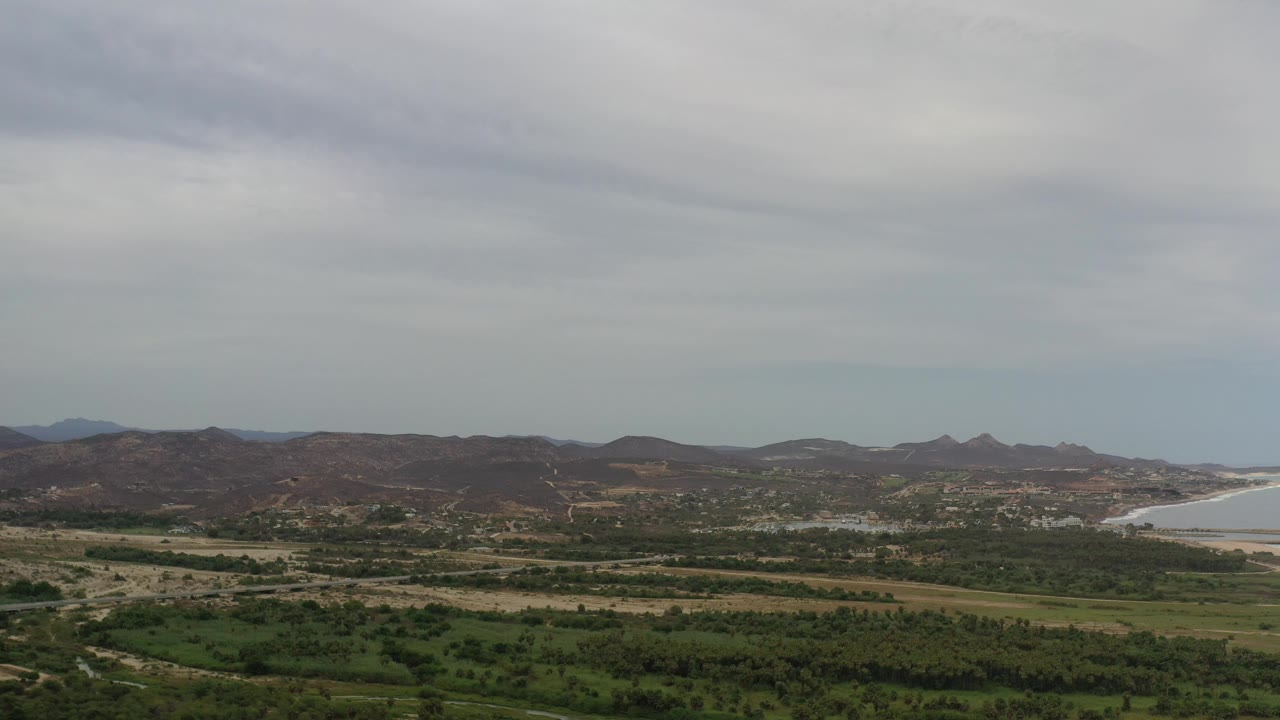 Coastal valley view with dark green vegetation, low hills, ocean, and overcast sky.