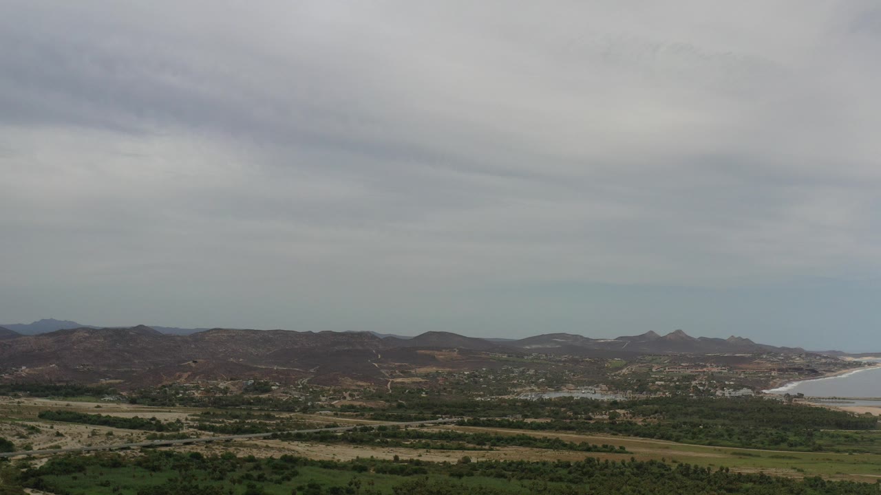 Elevated view of Los Cabos arid hills, green and yellow scrub, a distant shoreline on the right, and layered gray clouds, with no visible people.