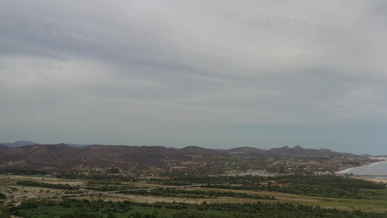 Wide Los Cabos landscape with a darker band of hills, green scrub across the valley floor, pale earth tracks, and a sliver of coast at right, with no visible people.