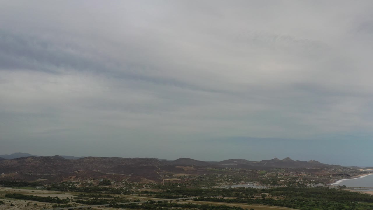 Wide aerial view of brown hills, green scrub, coastline, and gray clouds.