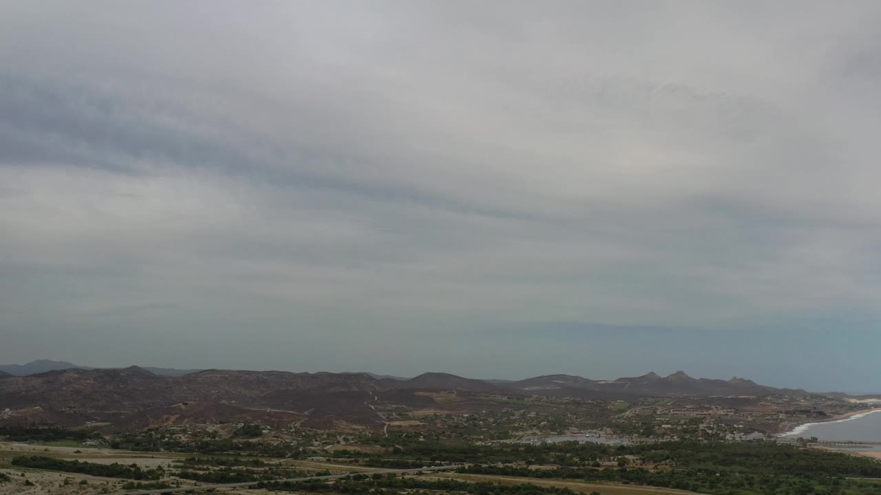 Los Cabos landscape frame with dry mountain ridges across the middle distance, green valley patches below, coastline to the right, and no visible people.