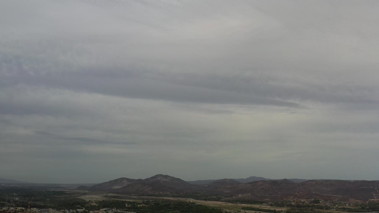 Aerial view in Los Cabos, Mexico with heavy gray clouds over low mountains, green terrain, scattered buildings, and no visible people.