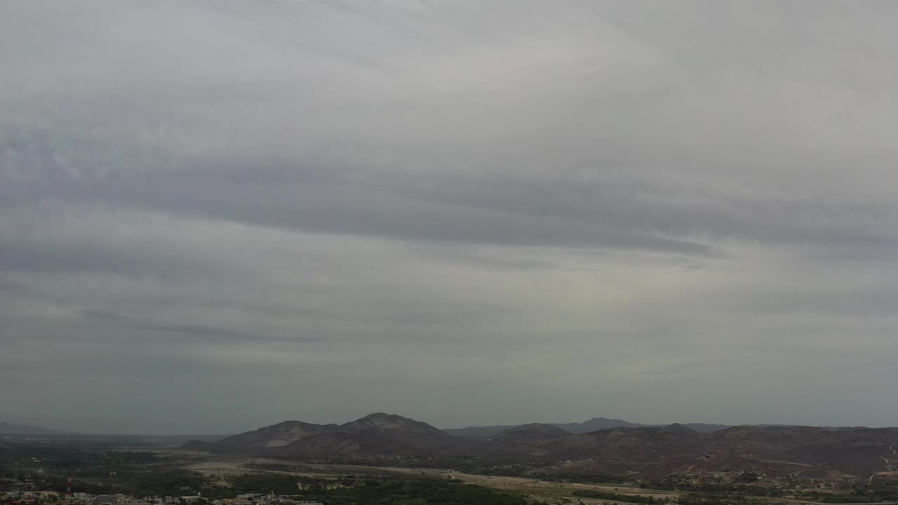 Wide Los Cabos aerial frame with a pale gray sky, dark cloud bands, low hills, green valley areas, distant buildings, and no visible people.