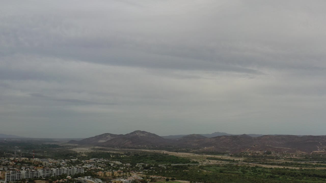 Opening frame of Los Cabos, Mexico, from high above, with a gray cloud layer over distant low mountains, green valley areas, and buildings along the bottom edge, no visible people.
