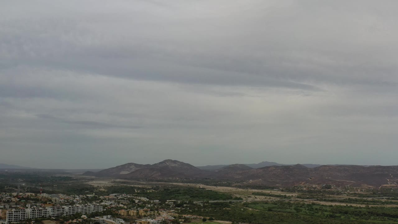 Los Cabos aerial frame with the mountain ridge centered farther back, a band of green terrain across the middle, and pale residential blocks near the lower foreground, no visible people.