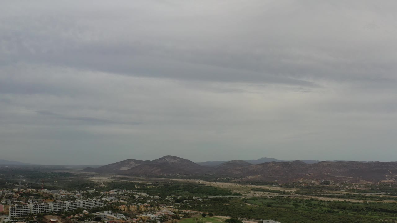 A high Los Cabos view shows layered gray clouds, brown-green hills across the horizon, a green valley band, and clustered buildings at the lower left, no visible people.