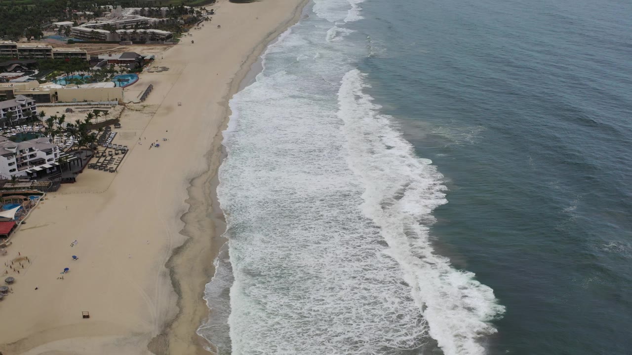 High aerial view over Los Cabos beach with white surf rolling along sand, beachfront buildings and pools at left, and tiny beachgoers visible on the open sand.